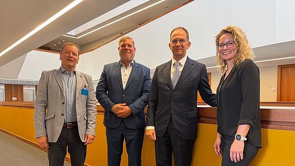 Drei Männer und eine Frau stehen in einer Reihe in einem Büroflur und blicken in Richtung Kamera
