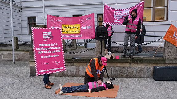 Fahrrad Demo Schwerin Eine Gruppe von Menschen ist am protestieren. Im Mittelpunkt übt jemand eine Herzdruckmassage an einer Puppe aus. Daneben ist ein Plakat zu sehen, auf dem steht: Wenn uns die TdL nicht langsam beatmet geht uns die Luft aus!