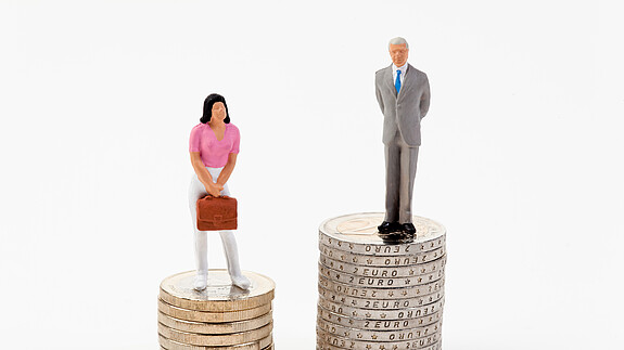Kreutz: „Der Arbeitsmarkt muss attraktiver sein als der Heiratsmarkt“ Das Foto zeigt eine männliche und eine weibliche Figur, die auf Münzstapeln stehen. Der Stapel der männlichen Figur ist deutlich höher.