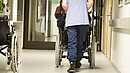 Eine Pflegekraft, von hinten zu sehen, schiebt einen älterne Menschen im Rollstuhl über einen Flur .