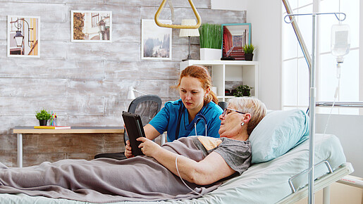 Pflege muss bezahlbar sein Eine ältere Frau liegt im Bett und eine Pflegerin hilft ihr dabei, ein Tablet zu bedienen
