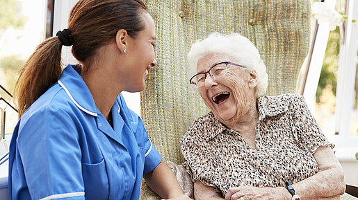 Szene aus Seniorenresidenz: Eine Seniorin sitzt auf einer Bank, neben ihr eine Pflegefachkraft. Beide lachen.