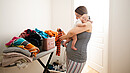Young mother holding sleepeing baby on her shoulder and trying to sort the laundry simultaniously. Mother combines everyday routines with raising her newborn baby.
